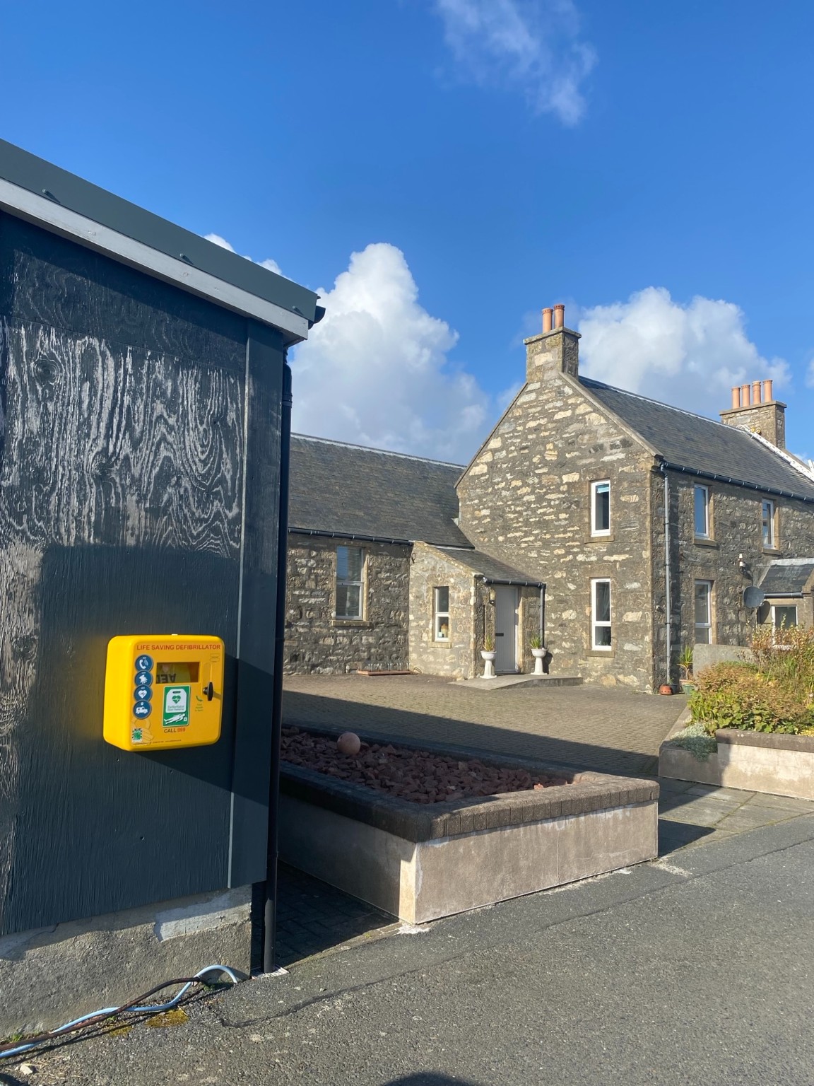 Yellow defibrillator cabinet outside the Aald Skül beside the main stone building