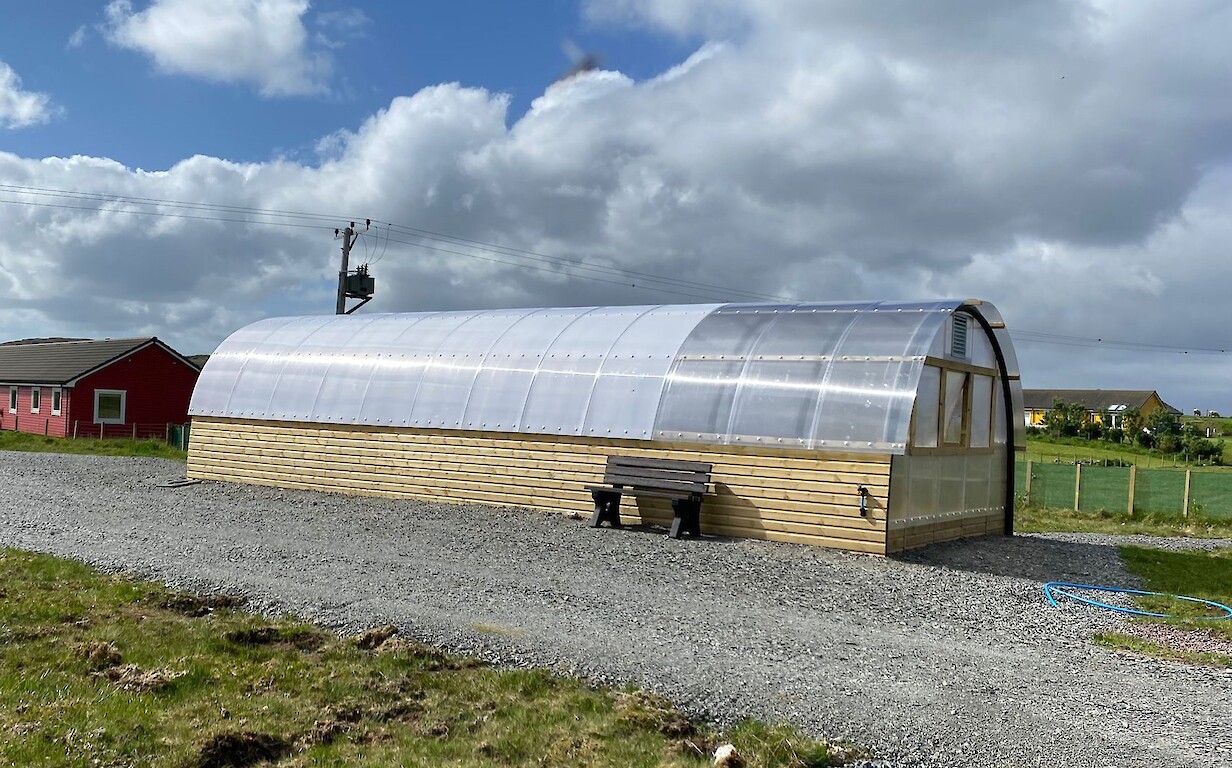 The big Polycrub at the Aald Sk&uuml;l, with timber sides, a translucent curved roof and a bench outside.